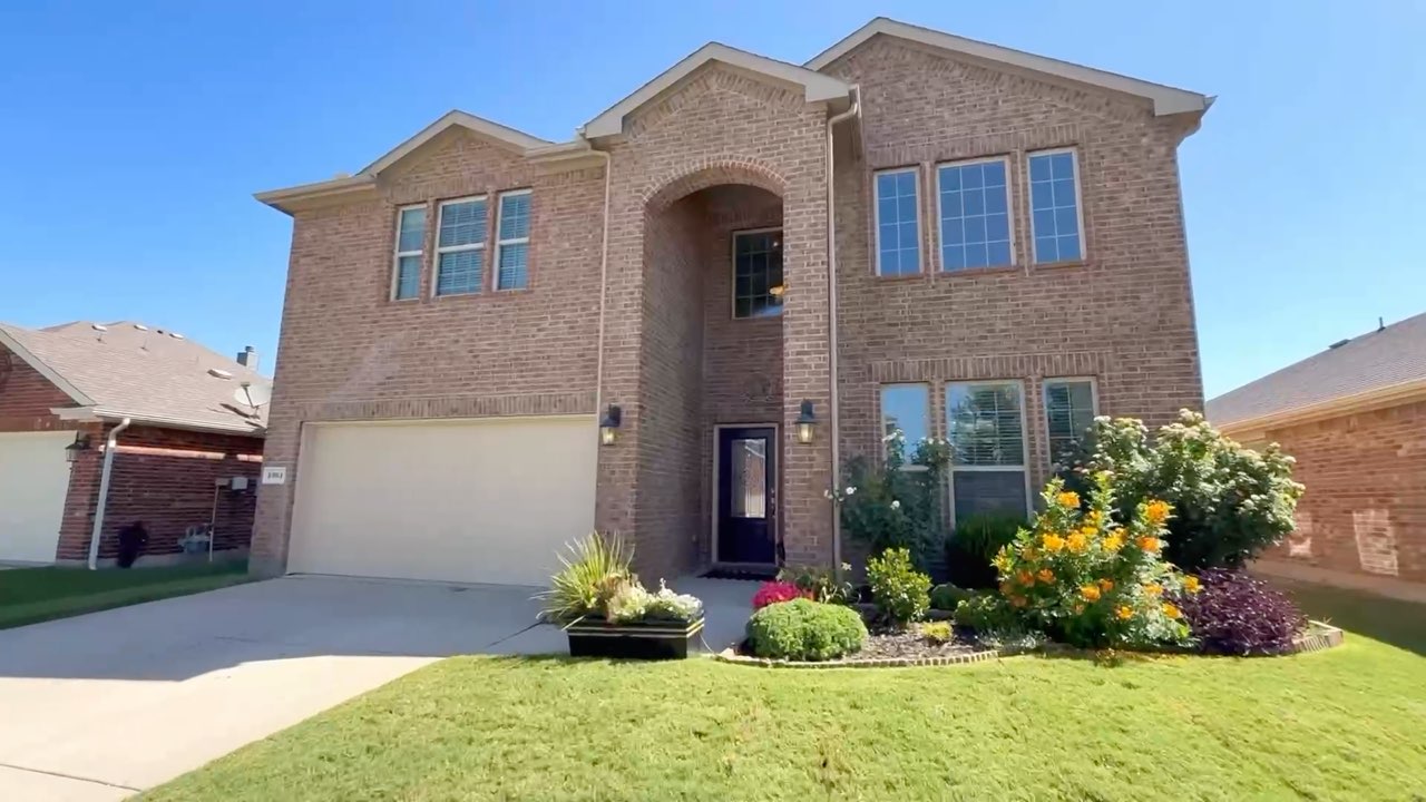 The two-story brick front of 1301 Coleman Dr in Villages of Melissa, with its flowerbeds and garage, from its listing film